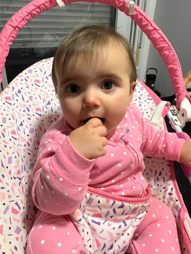 Joy na cadeirinha rosa, com o dedo na boca e olhos bem abertos.